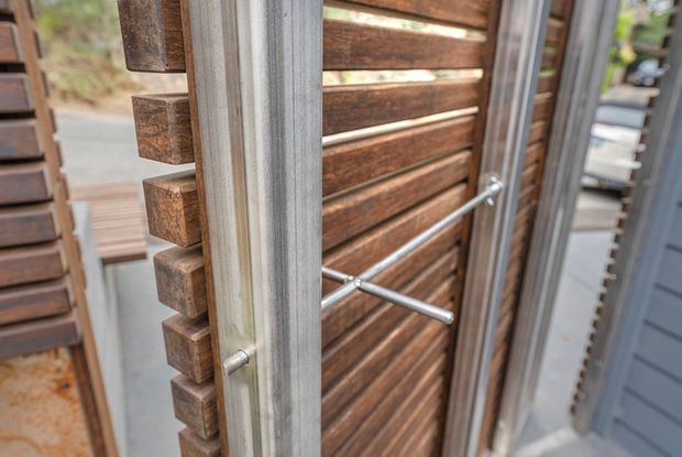 Close-up architectural detail of a timber slatted gate or screen with projecting square wood ends, framed by vertical metal members and fitted with diagonal metal bracing or hardware. Grey exterior cladding and a partially visible outdoor setting appear in the background.