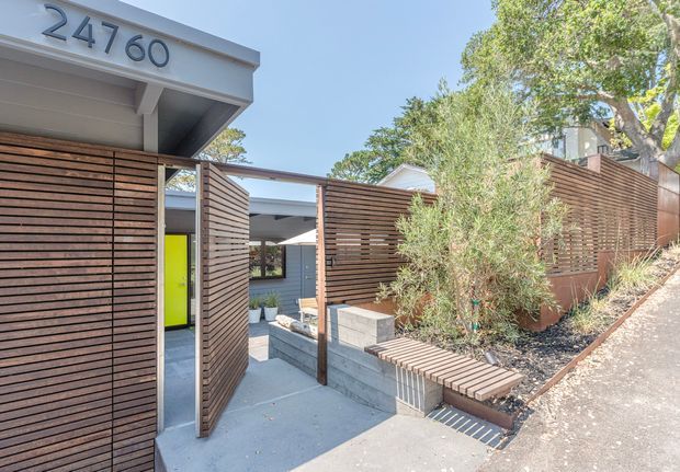 Street-level view of a contemporary residential entrance with the house number 24760 displayed on a projecting flat-roof canopy. Slatted timber privacy screens form the boundary and gate, opening onto a concrete path and stepped entry courtyard. The frontage includes dark grey masonry, a vivid lime-green entry door, built-in timber bench seating, and drought-tolerant planting on a sloped site.