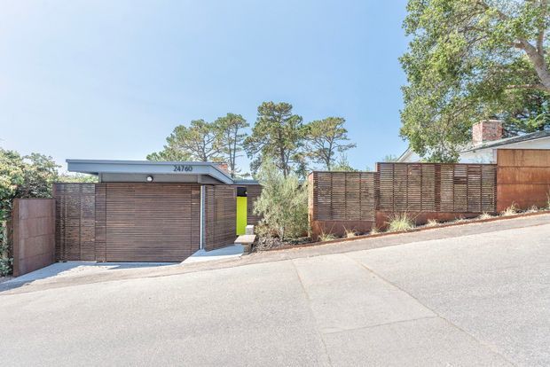 Street-level exterior view of a contemporary residential property on a sloping suburban site. The frontage features a wide timber-look slatted garage door, matching horizontal privacy screens and fencing, a low flat-roofed garage volume, and landscaped planting along the boundary. A timber-clad house volume and chimney are visible behind the fence.