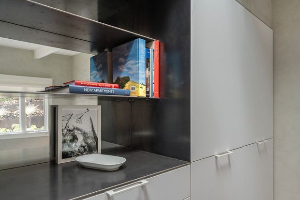 Close-up of a contemporary built-in storage and display unit beside a window. The unit combines glossy white cabinet doors and drawers with a dark timber or black panelled backing, open shelving containing books and artwork, and a dark countertop surface. The compact composition has clean lines and minimal detailing.