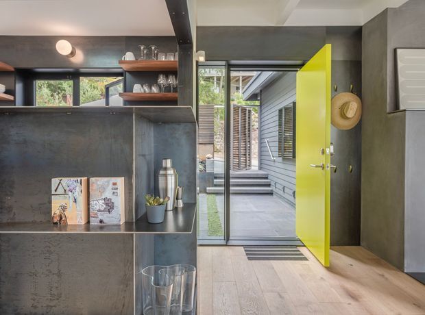 A modern interior entryway with a dark textured built-in shelving and bar-like joinery, displaying glassware and small decor. A vivid lime-yellow pivot door opens to a covered outdoor walkway and steps. The space combines grey concrete-like surfaces, timber flooring, black-framed glazing, and contemporary architectural detailing.