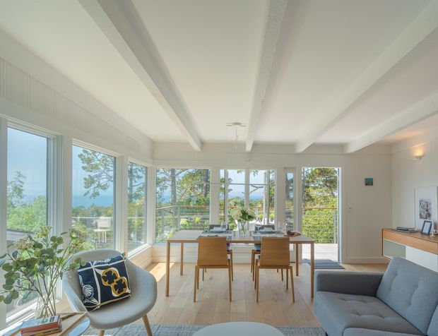 Bright, airy open-plan living and dining room with extensive windows overlooking greenery and distant water or landscape. The room features a pale timber floor, white walls and ceiling with exposed boxed ceiling beams, a light timber dining table and chairs, a grey sofa, a sculptural lounge chair, and minimal contemporary furnishings. A built-in sideboard and framed artwork sit along the right wall, while plants and blue decorative accents add colour.