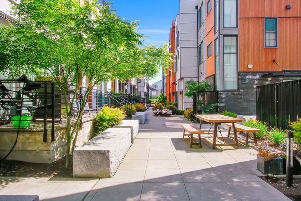 A bright landscaped pedestrian courtyard or shared walkway between contemporary multi-storey residential buildings. The space features large-format paved surfaces, raised garden beds, mature trees, ornamental grasses, concrete seating blocks, black metal fencing, and a timber picnic table with benches. The surrounding buildings combine warm timber cladding, grey panels, and extensive vertical glazing.