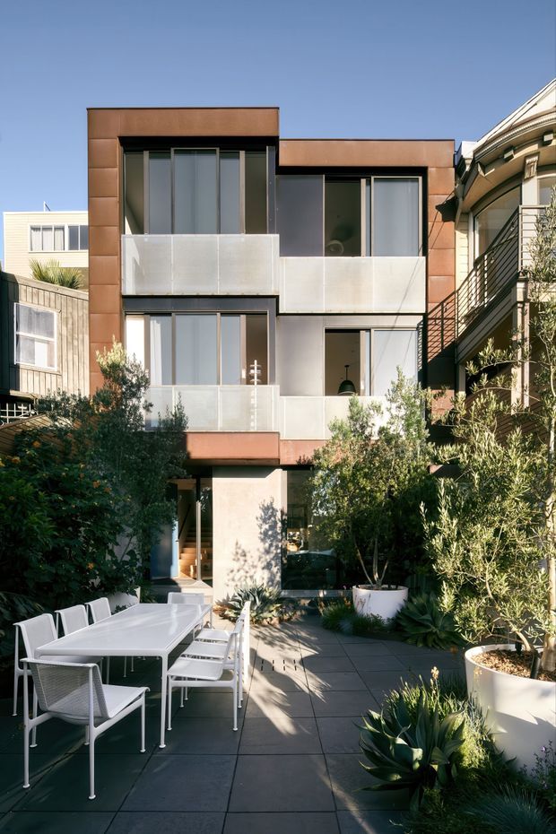 A contemporary multi-storey urban residence viewed from a landscaped outdoor terrace. The façade combines dark metal-framed glazing, pale rendered panels, and warm brown cladding, with translucent balcony balustrades and projecting box forms. The terrace is paved with large square slabs and furnished with a white outdoor dining table and chairs, surrounded by dense greenery and potted plants. Adjacent residential buildings are visible on both sides.