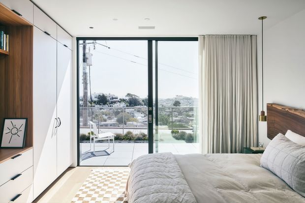 Bright contemporary bedroom with a low-profile bed, white bedding, integrated white wardrobe cabinetry and warm timber shelving. Full-height glazed sliding doors open to a balcony, with floor-to-ceiling curtains and an urban suburban view beyond. The room has pale timber flooring, a small patterned rug, and a simple suspended bedside light.