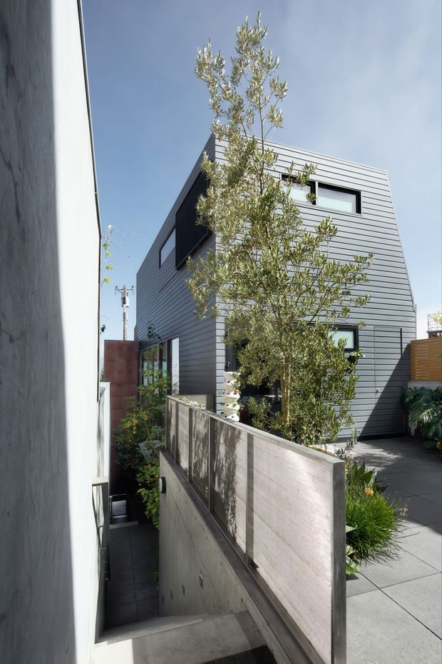 Exterior view of a compact modern multi-storey home in a narrow urban setting. The building has horizontal grey cladding, dark-framed rectangular windows, a flat roofline, and a paved terrace or courtyard with raised concrete planters and established planting. A narrow stair passage runs between the neighbouring wall and the house.