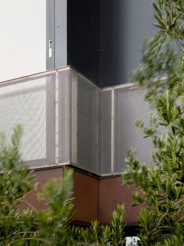 Close-up exterior architectural detail of a contemporary building corner, featuring dark and light vertical wall panels above perforated metal screening or balustrade panels. Dense green planting partially obscures the lower facade, creating a layered urban architectural composition.