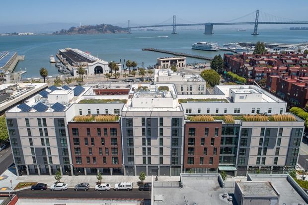 Aerial view of a contemporary multi-storey apartment building with flat roofs, rooftop planting, and a mixed white, grey, and red-brick facade. The building overlooks a waterfront with piers and ferries, with a large suspension bridge visible in the background.