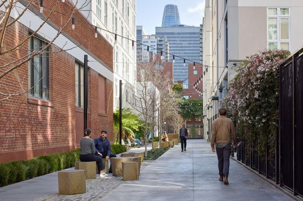 A landscaped pedestrian laneway in a dense urban setting, bordered by red-brick and rendered multi-storey buildings. The walkway includes concrete paving, low planting, bare deciduous trees, seating blocks, and black metal fencing, with modern high-rise buildings visible in the distance.