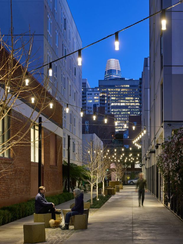 An urban pedestrian laneway at dusk, framed by multi-storey contemporary buildings. String lights are suspended overhead and wrapped around leafless trees, illuminating concrete paving, planted edges, and simple block seating where people are gathered. A brightly lit commercial tower rises in the background, creating a lively, intimate public-space atmosphere.