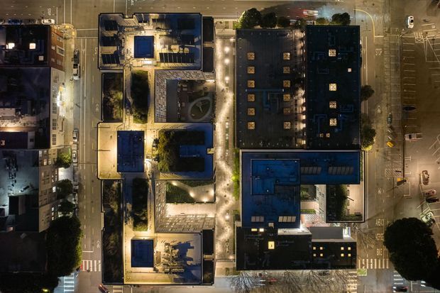 Aerial nighttime view of a dense urban block with several flat-roofed buildings, rooftop courtyards and terraces, landscaped paths, surrounding streets, marked pedestrian crossings, and an adjacent parking area. Warm architectural and pathway lighting contrasts with dark roofs and blue-toned rooftop surfaces.