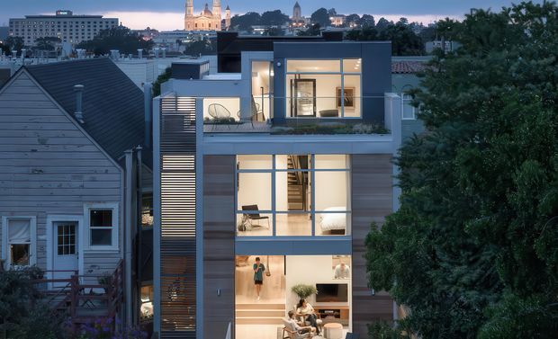 Twilight exterior view of a narrow, multi-storey contemporary urban residence. The house features a strong vertical composition, large floor-to-ceiling glazing, dark and timber-toned facade panels, a rooftop terrace, and projecting upper-level volumes. Older neighbouring homes and a city skyline with illuminated civic buildings are visible in the background.