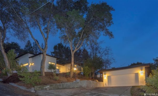 Twilight exterior view of a modern multi-level residence set among mature trees on a sloped site. The house has smooth light-coloured walls, dark low-pitched roof planes, illuminated rectangular windows, retaining walls, and a separate attached-looking garage structure with a wide garage door and concrete driveway.