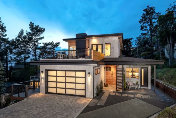 Twilight exterior photograph of a contemporary multi-level hillside home. The house has a flat and low-pitched roof composition, rendered light walls, dark trim, extensive timber cladding, a large glazed garage door, upper-level balcony, and a covered entry terrace. Tall trees and a sloping site surround the dwelling.