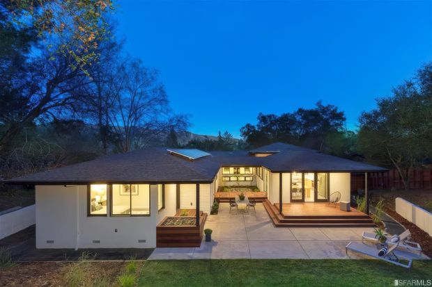 Twilight exterior view of a single-storey residential home with a low-pitched roof, broad glazed openings, and a central courtyard-style patio. The house has light rendered walls, dark roof tiles, timber decking, concrete paving, raised garden beds, and warm interior lighting, set among mature trees.