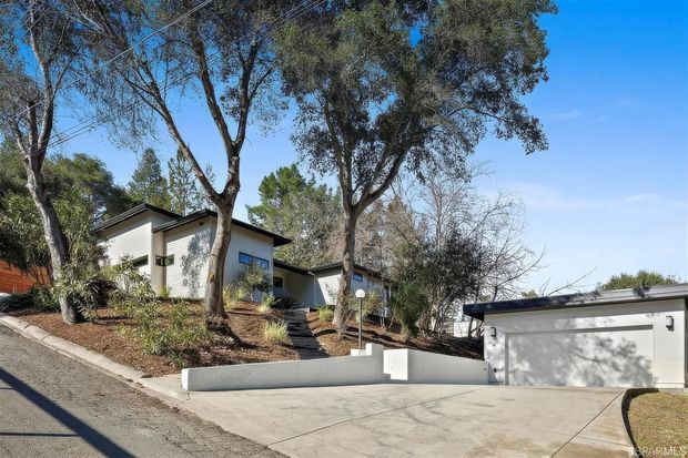 Exterior view of a modern single-storey residence on a sloped, landscaped site, with white rendered walls, dark low-pitched rooflines, large windows, and a detached or projecting double garage accessed by a broad concrete driveway. Mature trees and planted garden beds surround the house.