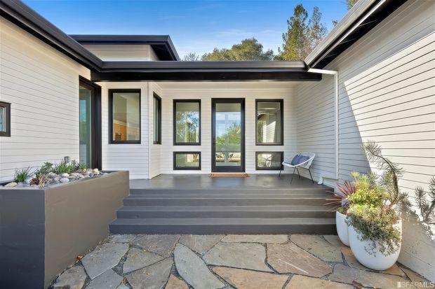 A contemporary single-storey courtyard entry with white horizontal cladding, crisp black-framed windows and doors, a low-pitched roofline, and broad dark steps leading to a covered entrance. Irregular natural stone paving, raised planters, and potted landscaping soften the modern facade.