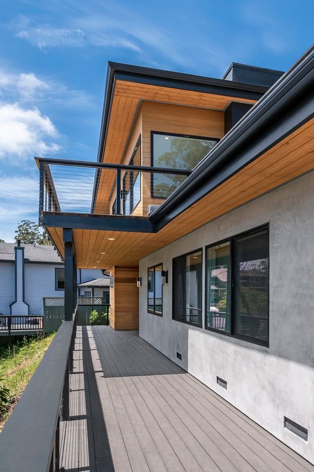 Exterior view of a contemporary multi-level home showing a long elevated timber deck, black metal balustrades, projecting roof planes, timber soffits and cladding, dark-framed glazing, and a textured light grey wall finish. The neighbouring suburban houses are visible in the background.