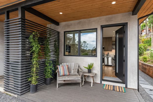 Covered front entry porch with a timber-lined ceiling, exposed dark structural beams, grey rendered walls, charcoal horizontal privacy screening, and a small outdoor seating area with potted plants. A glazed door opens into a kitchen, while the deck and garden retaining wall frame the entrance.