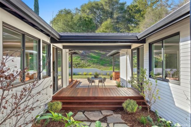 A bright contemporary courtyard connecting two wings of a low-rise home. The covered outdoor deck features timber decking, steps, a small dining setting, large glazed openings, and light horizontal exterior cladding. Native-style planting and a landscaped path frame views toward a green hillside and water beyond.
