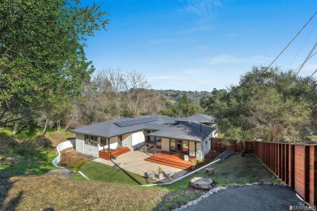 Elevated exterior view of a low-slung single-storey home set within a wooded, hilly landscape. The house has dark grey low-pitched roofs, light rendered walls, extensive glazing, warm timber decking and a paved central terrace. Solar panels are visible on the roof, with a red-brown timber boundary fence and a curved concrete path around the property.
