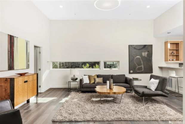 Bright contemporary living room with white walls and ceiling, recessed lighting, grey wood-look flooring, a large textured area rug, dark grey sofa and armchair, and a round timber coffee table. Built-in shelving and a compact kitchen or bar area are visible at right, while a timber sideboard and abstract artwork add warmth and contrast.