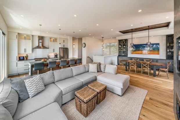 Bright open-plan interior combining a contemporary kitchen, living room and dining/bar area. The space features pale built-in cabinetry, stainless-steel appliances, a large light-grey sectional sofa, warm timber flooring, a timber dining table, and a linear wood ceiling feature above the dining zone. Recessed lighting, pendant lights, and abundant glazing create an airy, comfortable family entertaining space.