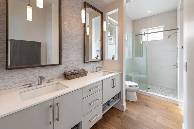 Bright contemporary bathroom with a long double vanity, white countertop, pale grey cabinetry, large framed mirrors, textured light wall tile, and warm timber flooring. A glass-enclosed shower and toilet are visible in the adjacent section, with simple pendant and recessed lighting.