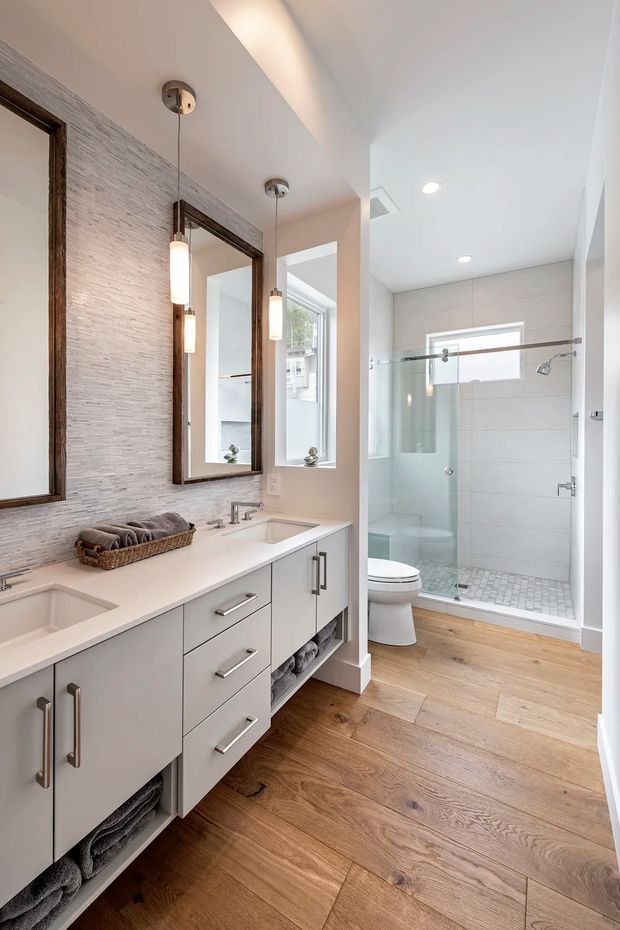 Bright contemporary bathroom with a long double vanity, pale grey cabinetry, white benchtop, twin undermount sinks, and large framed mirrors. A glass-enclosed walk-in shower and toilet occupy the rear, with pale tiled walls and a small mosaic shower floor. Warm timber-look flooring, recessed ceiling lights, and slim pendant lights create a clean, refined finish.