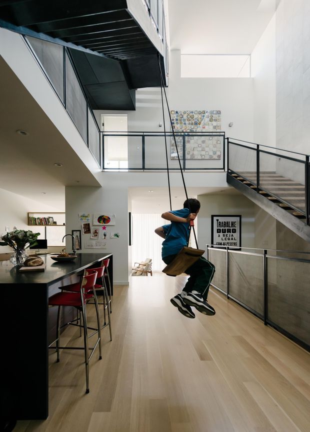 A bright, open-plan multi-level residential interior with a kitchen and breakfast island in the foreground, a large pale timber floor, black-framed glass balustrades, and a dramatic suspended metal stair and mezzanine structure. A child swings through the central double-height space. White walls, abundant glazing, contemporary artwork, and black-and-red bar stools create a lively contemporary atmosphere.