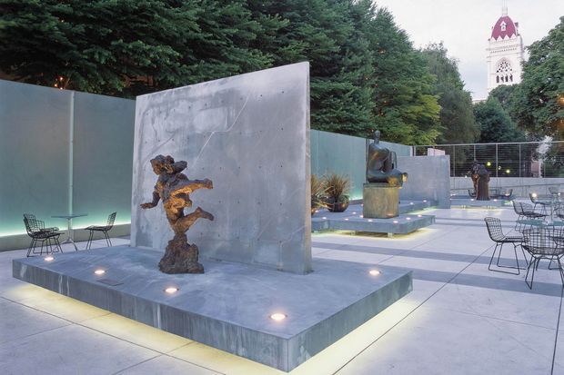 A landscaped urban courtyard or public plaza with large concrete architectural screens and raised plinths displaying sculptural artworks. The paved space includes minimalist metal café tables and chairs, integrated ground lighting, planting, and a church tower visible beyond the trees.