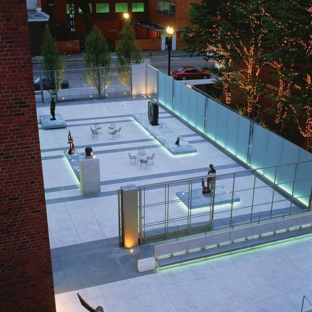 Elevated view of a contemporary urban outdoor plaza or terrace beside a brick building, featuring pale tiled paving, glass and metal balustrades, sculptural artworks, small café-style tables and chairs, illuminated benches and low linear lighting. Trees wrapped in warm string lights and surrounding city buildings create an evening atmosphere.