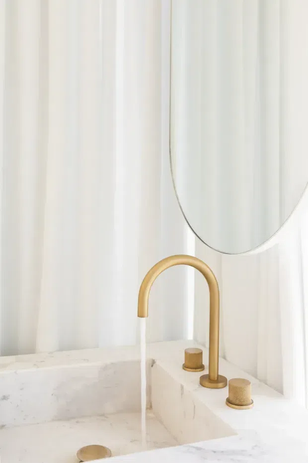 Close-up view of a refined bathroom vanity featuring a white marble or stone basin and benchtop, a tall curved brushed-gold tap, matching cylindrical controls, and a round mirror. Sheer white curtains provide a soft, bright backdrop.