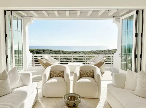 Bright coastal living room opening through fully glazed bi-fold doors onto a deck or balcony with ocean views. The room features white upholstered sofas and sculptural armchairs, a pale textured rug, a low coffee table, a white exposed-beam ceiling, and a horizontal metal balustrade outside. The overall design is airy, refined, and monochromatic with strong indoor-outdoor connection.