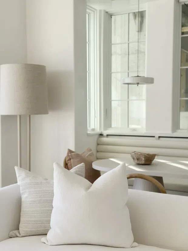 A bright, softly styled sitting and dining area with a predominantly white and cream palette. The foreground features a light upholstered sofa with textured cushions, while a round dining table and curved wooden chair sit by tall multi-pane windows. A built-in window seat with horizontal padded upholstery, a floor lamp, and a suspended circular pendant complete the calm, minimalist interior.