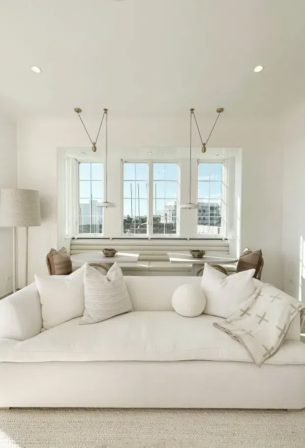 Bright, airy living room with a large white upholstered sofa in the foreground, layered neutral cushions, a textured throw, and a pale woven rug. Behind it is a built-in window seat beneath a bank of tall divided-light windows, flanked by slim pendant lights and recessed ceiling lights. The room has a calm, monochromatic neutral palette with abundant natural light and understated coastal styling.