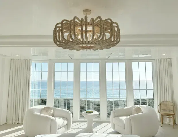 Bright coastal living room with large white upholstered armchairs, a small pedestal side table, full-height multi-pane windows overlooking the ocean, and sheer white curtains. A prominent woven rattan or rope chandelier hangs from the ceiling, while sunlight creates strong geometric shadows across the pale floor.