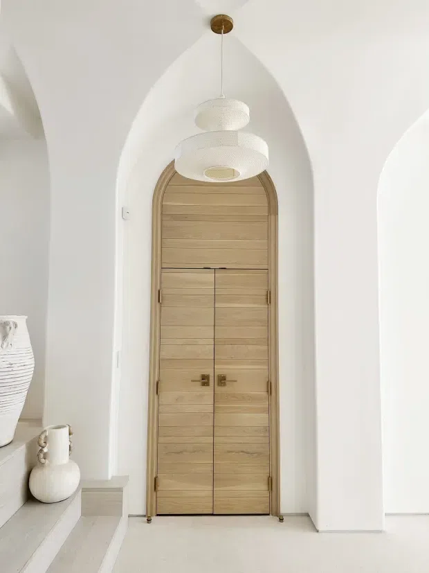A bright, minimalist interior passage with a tall arched doorway made from horizontal light timber boards, set within sculpted white plaster arches. Two white pendant lights hang in front of the door, while pale flooring and a small built-in ledge with ceramic vessels reinforce the calm, refined palette.