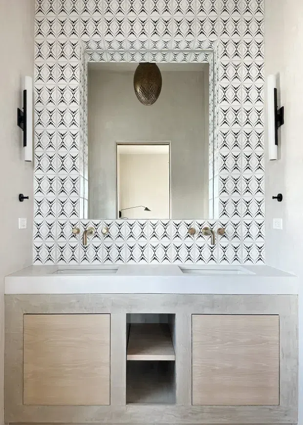 A contemporary bathroom vanity wall featuring a wide double basin with wall-mounted brass fixtures, a large framed mirror, and a highly patterned black-and-white tiled splashback. The vanity is clad in a pale textured finish with light timber cabinet fronts and an open central shelf. Minimal black wall sconces and door hardware contrast with the predominantly white, grey, and warm wood palette.