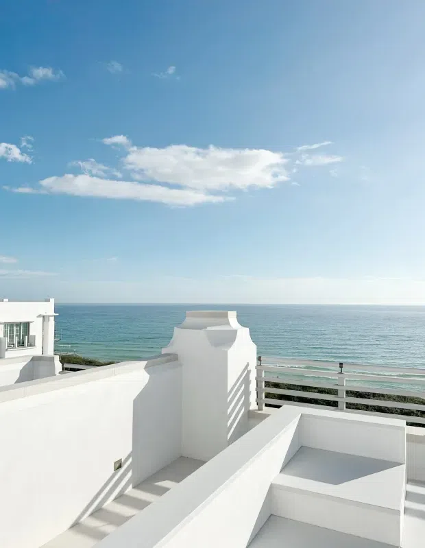 Bright coastal rooftop terrace overlooking the ocean, with white rendered parapet walls, sculptural curved architectural detailing, stepped levels, and horizontal metal balustrades. The scene has a clean, sunlit Mediterranean-inspired character with expansive sea and sky views.