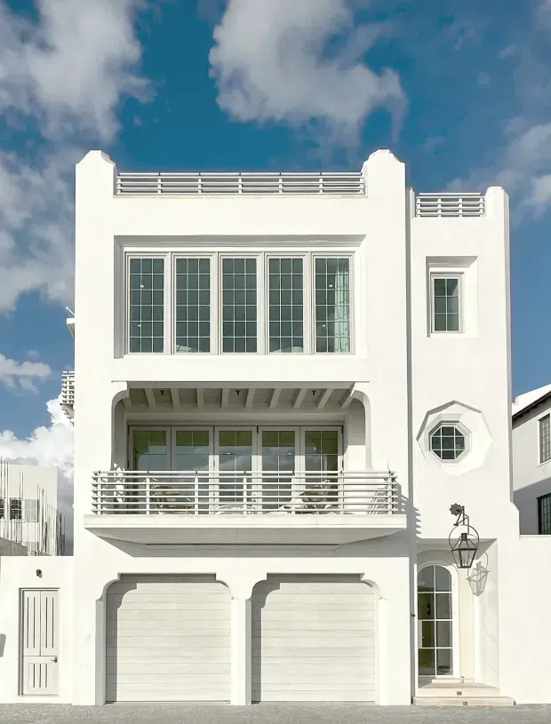 Bright daytime photograph of a narrow, multi-storey white rendered building facade. The elevation features two garage doors at ground level, a tall glazed entrance, large multi-pane windows, projecting balconies with horizontal metal railings, rooftop terraces, arched detailing, and a small octagonal window. The composition has strong geometric symmetry and a crisp white-and-grey palette against a blue sky.