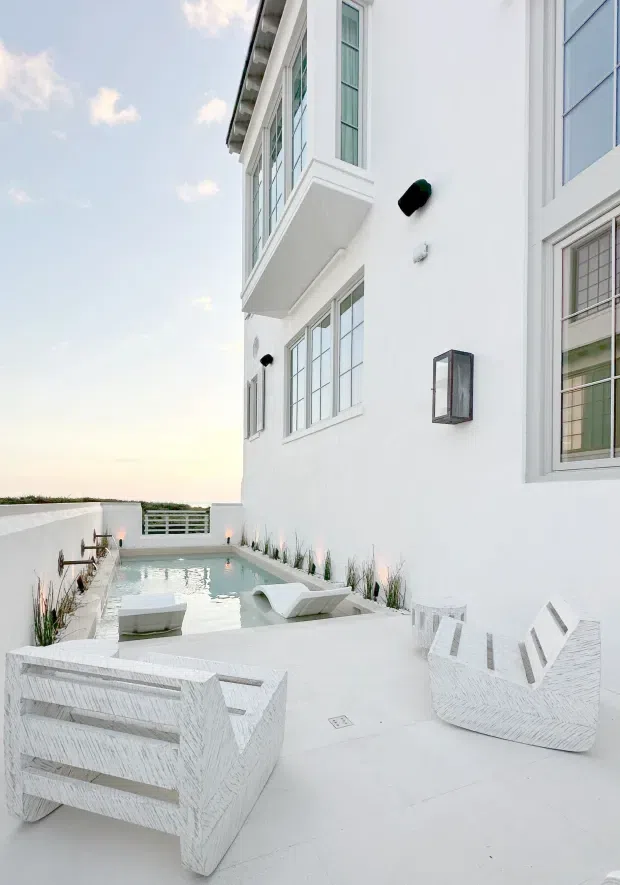 A bright white exterior terrace beside a multi-storey house, featuring a narrow rectangular swimming pool, white sculptural outdoor lounge chairs, integrated linear planting, white rendered walls, and tall gridded windows. The terrace has an airy, minimalist resort-like appearance with soft daylight and a distant horizon visible beyond the boundary wall.