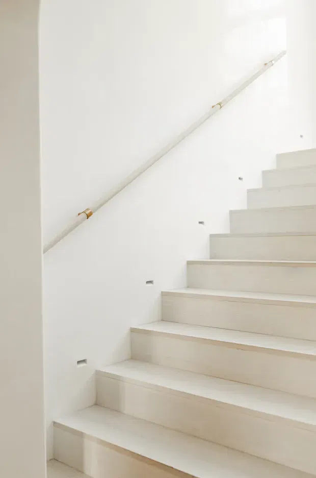 Bright minimalist interior staircase with pale, light-toned tiled or stone-clad treads and risers, white walls, recessed step lighting, and a slim white handrail with small brass or gold-toned fittings. The stairway rises diagonally through a clean, uncluttered space.