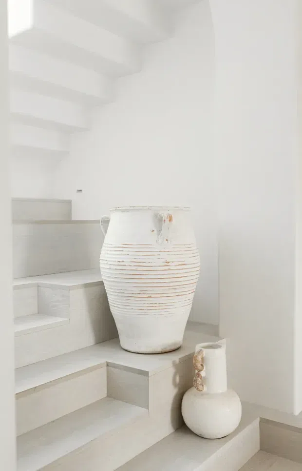 A softly lit minimalist stairway with broad pale stone or tile steps and crisp white walls. Two large white sculptural ceramic vessels with subtle horizontal ridges and organic forms are displayed on the stair landing, creating a calm, monochromatic composition. The stairwell has a high, clean-lined ceiling with simple projecting beams or steps visible overhead.