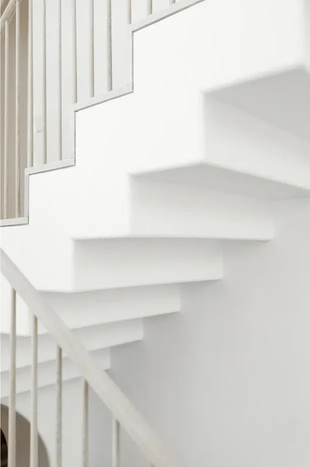 Close-up view of a minimalist white staircase with thick floating-style treads, a white structural wall, and simple vertical white balusters. The image is tightly framed around the stair geometry and railing, with a bright, monochromatic architectural interior.