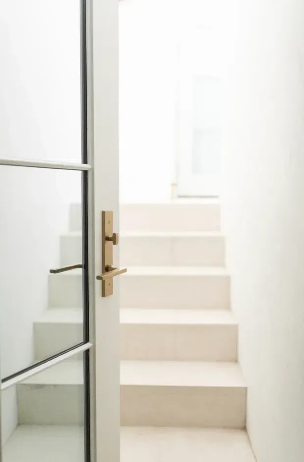 Bright minimalist stairway viewed through a partially open glass-panel door. The narrow staircase has pale steps and white walls, with a slim dark-framed glazed door and brushed brass or bronze hardware in the foreground. A reflected view of the stair and door appears in the glazing.