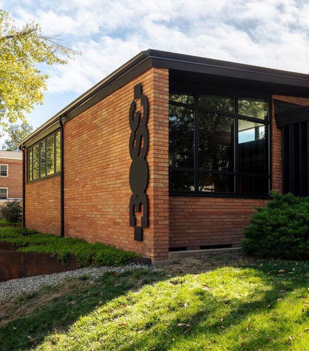 Exterior view of a low-rise brick building with a contemporary flat roof, dark metal-framed glazing, and a prominent vertical sculptural sign or artwork mounted on the brick facade. The building sits on a landscaped, slightly sloping grass site with shrubs and mature trees nearby.