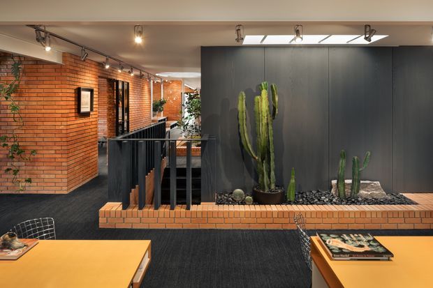 A stylish commercial interior with exposed red brick walls, dark charcoal wall panelling, black carpet, and a central raised planter filled with tall cacti and succulents. Black metal railings and stair openings define the circulation area, while warm timber tables and track lighting create an inviting, contemporary hospitality or workplace atmosphere.