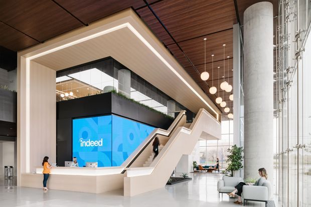 A spacious contemporary commercial office lobby with a dramatic sculptural staircase, large digital display, reception counter, exposed cylindrical structural columns, full-height glazing, and a warm timber-lined ceiling with integrated linear lighting. Pendant globe lights hang over a lounge and informal meeting area, while greenery and pale upholstered seating soften the polished, high-volume interior.