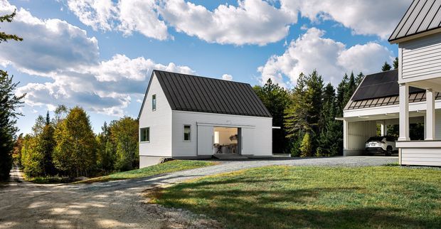 Exterior view of a contemporary white-clad residential property in a wooded setting. The main detached building has a simple gable form, vertical black standing-seam metal roofing, white horizontal cladding, and large glazed openings. A covered carport or verandah structure is visible on the right, with a gravel driveway and lawn in the foreground.